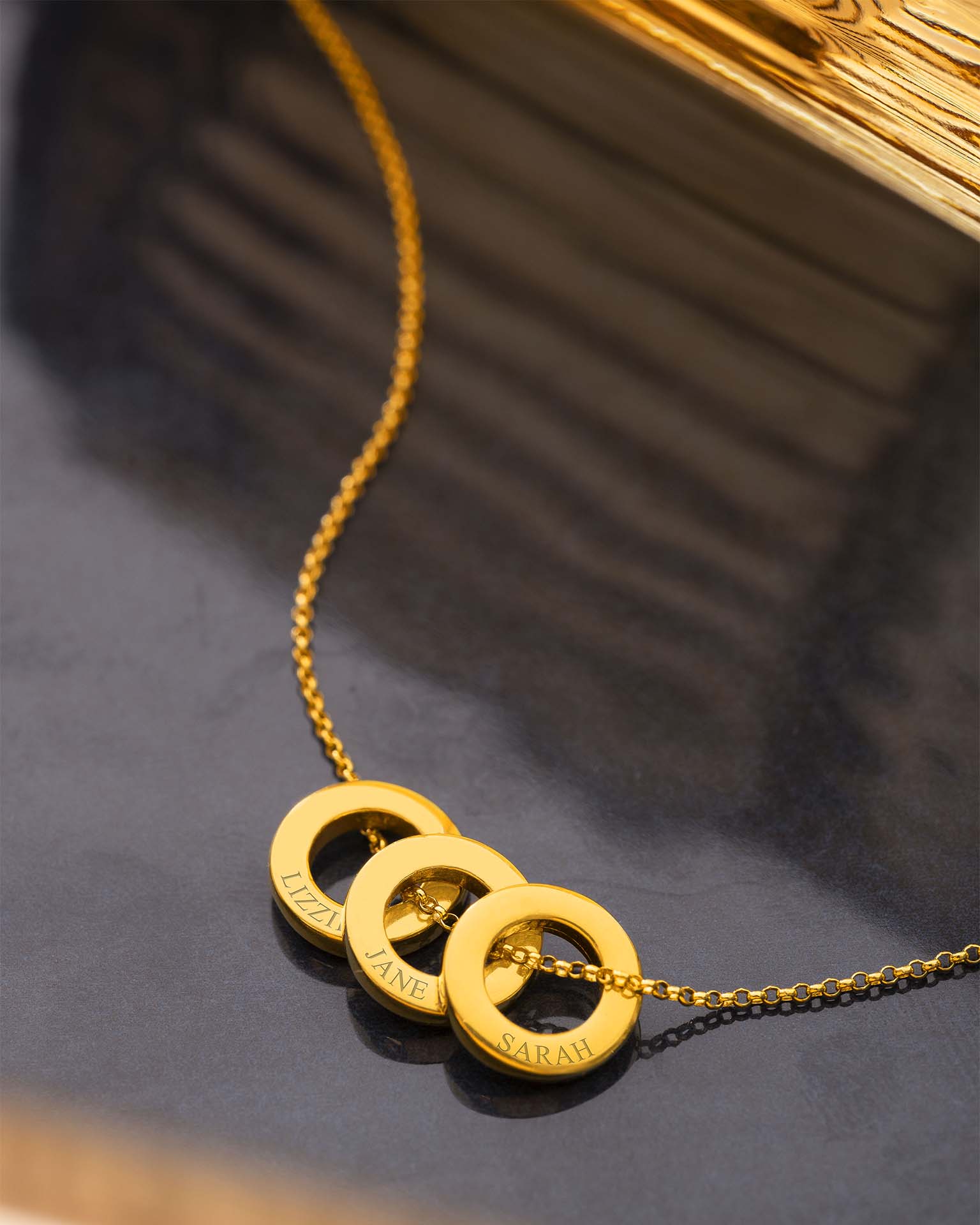 Gold necklace with three interlocking rings engraved with names on a dark surface.