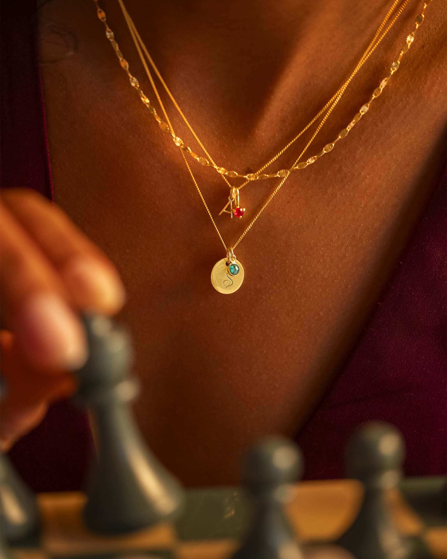 Close-up of a person wearing gold necklaces with small pendants on a blurred chessboard background.