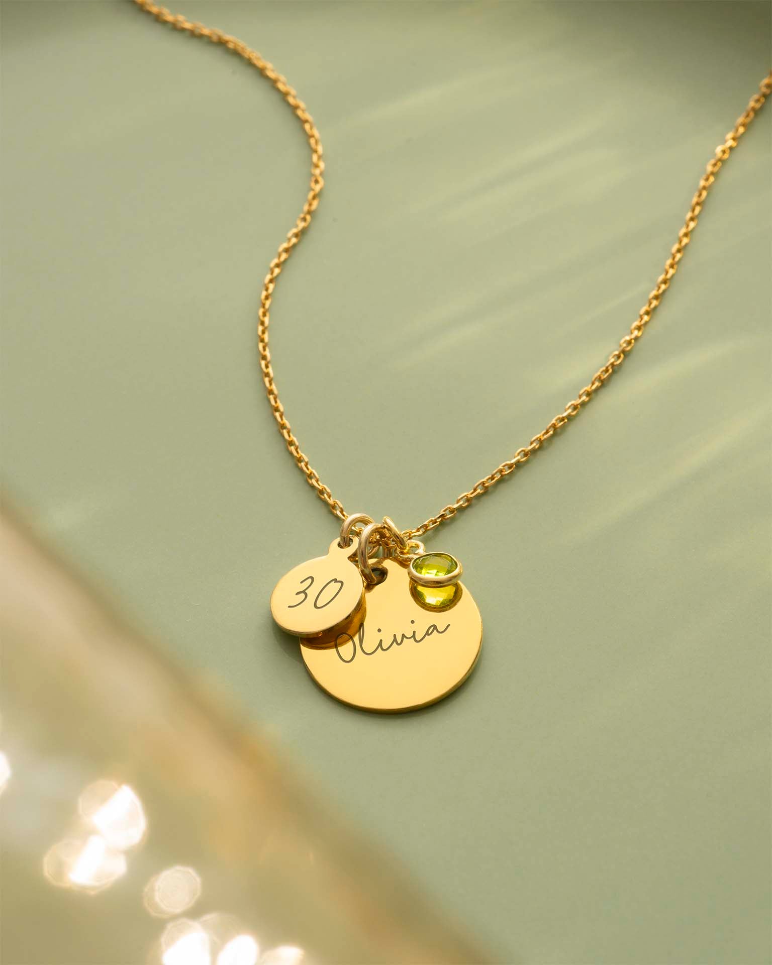 Gold necklace with two round pendants and a green gemstone on a light green background