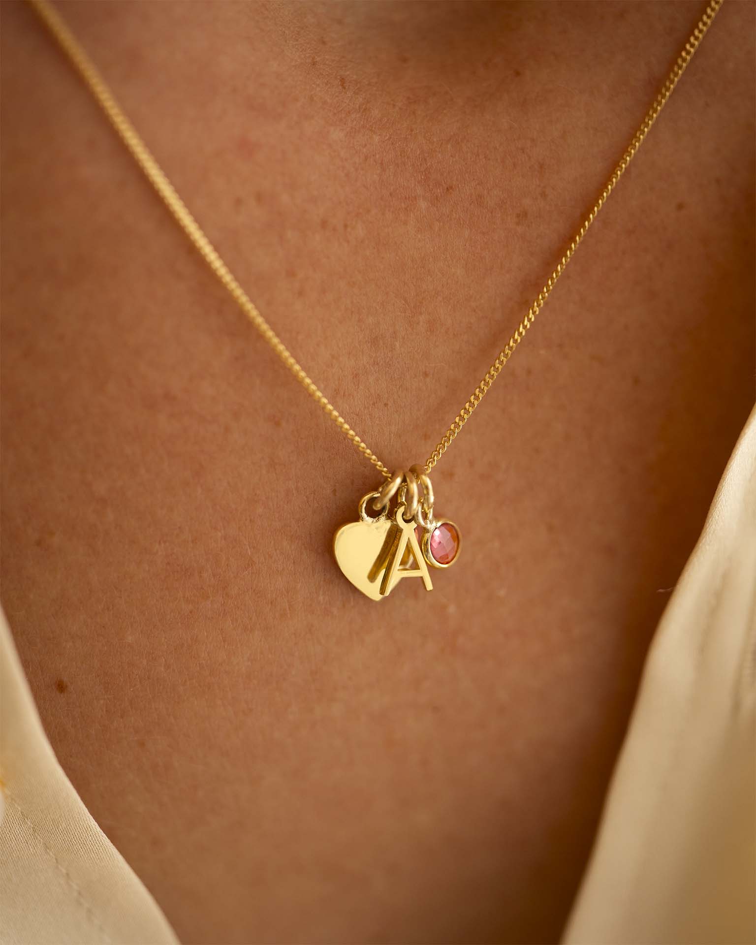 Gold necklace with heart, letter 'A', and pink gemstone pendant on a person's neck. 