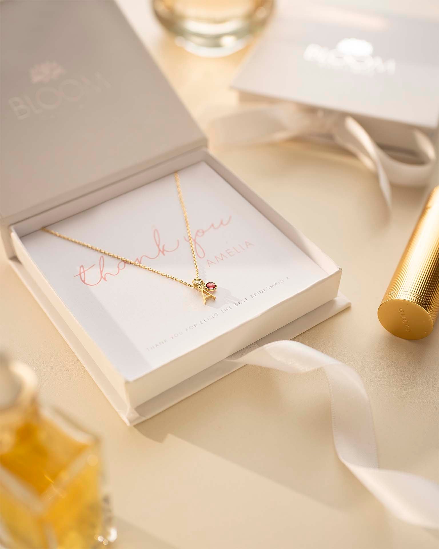 Gold necklace with a small round pendant on a branded jewelry box with a white ribbon and perfume bottle in the background.