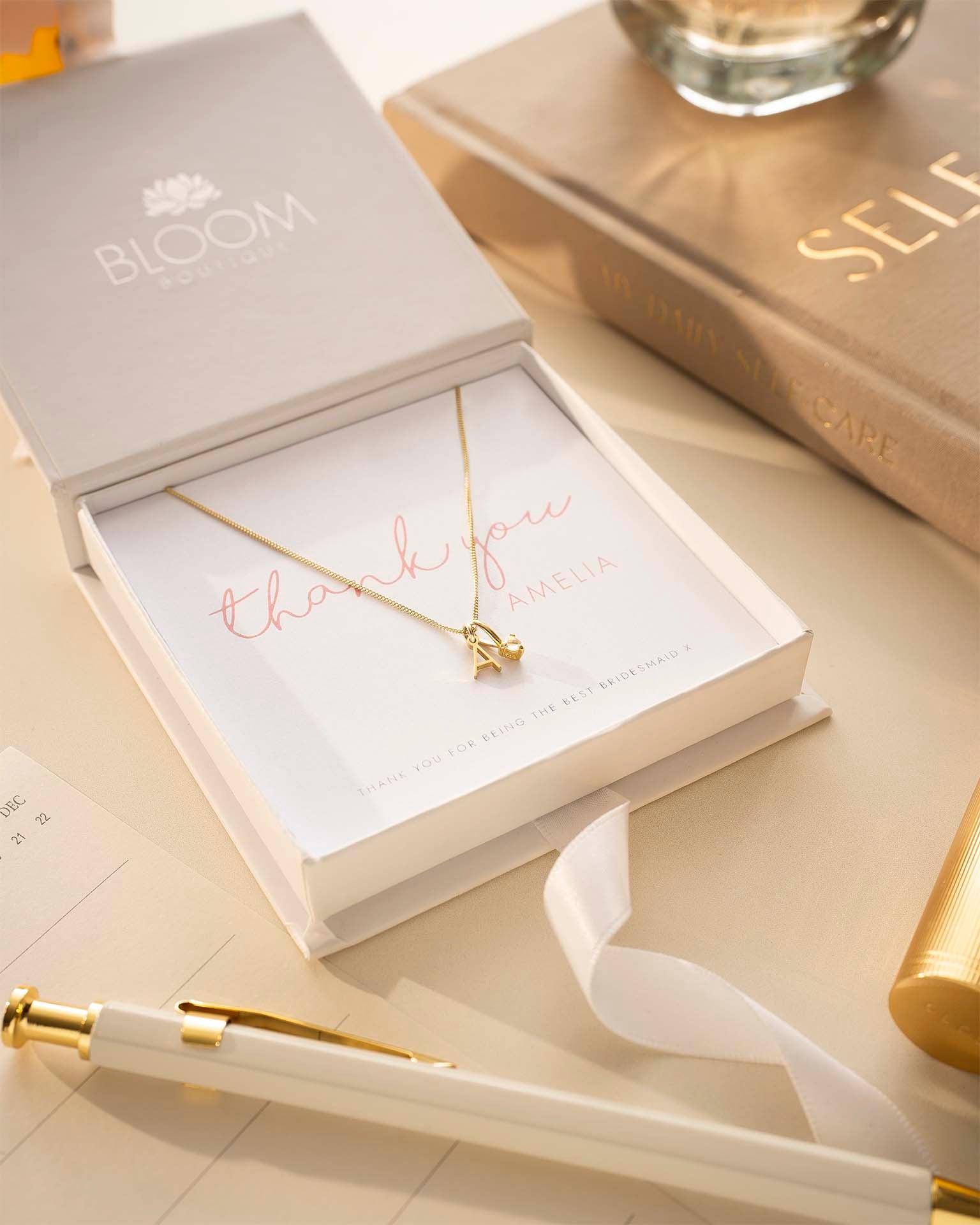 Gold necklace in a 'BLOOM' box with a pen and book on a light surface