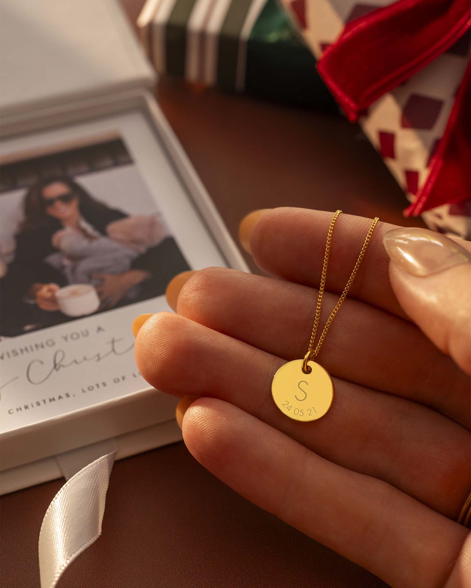 Gold necklace with a round pendant held by a hand, with a blurred background of a gift and photo album.