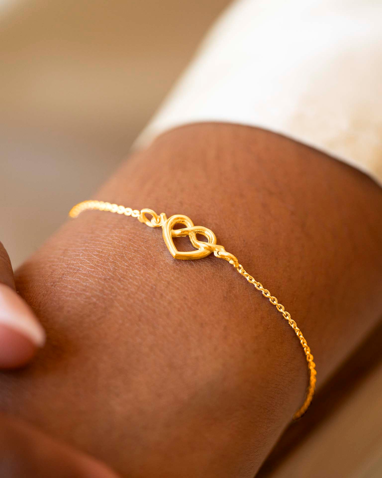 Gold bracelet with a heart and infinity symbol on a wrist against a neutral background