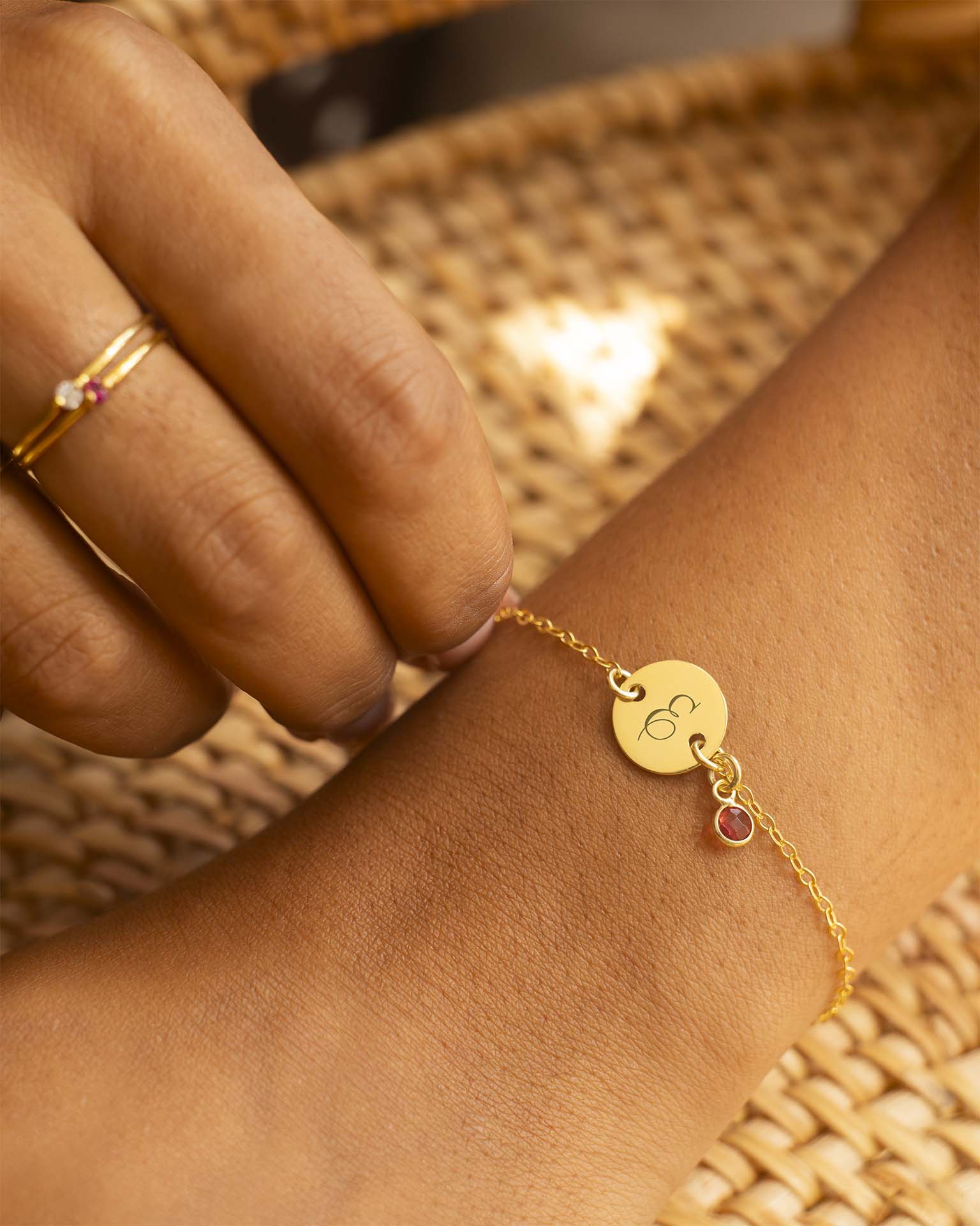 Gold bracelet with a round tag and small round gemstone on a wrist against a woven background