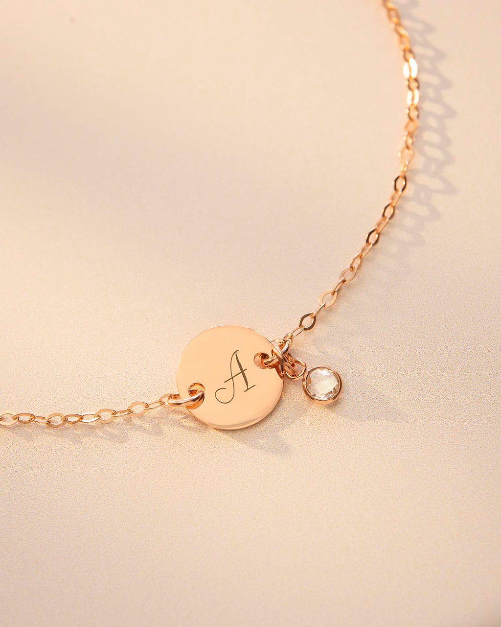Gold bracelet with a round disc charm featuring an 'A' and a small round charm on a beige background