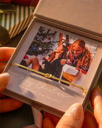 Person holding a Christmas card with two people and a tree, inside an open box. | Christmas