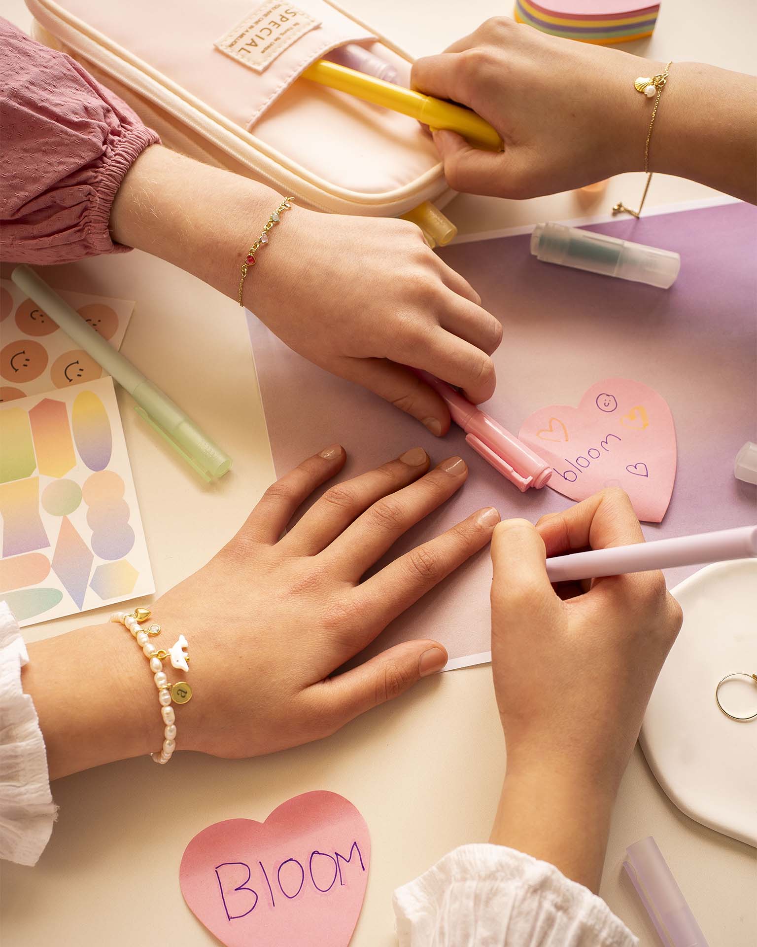 Two people decorating pink heart-shaped cards with markers on a light surface.