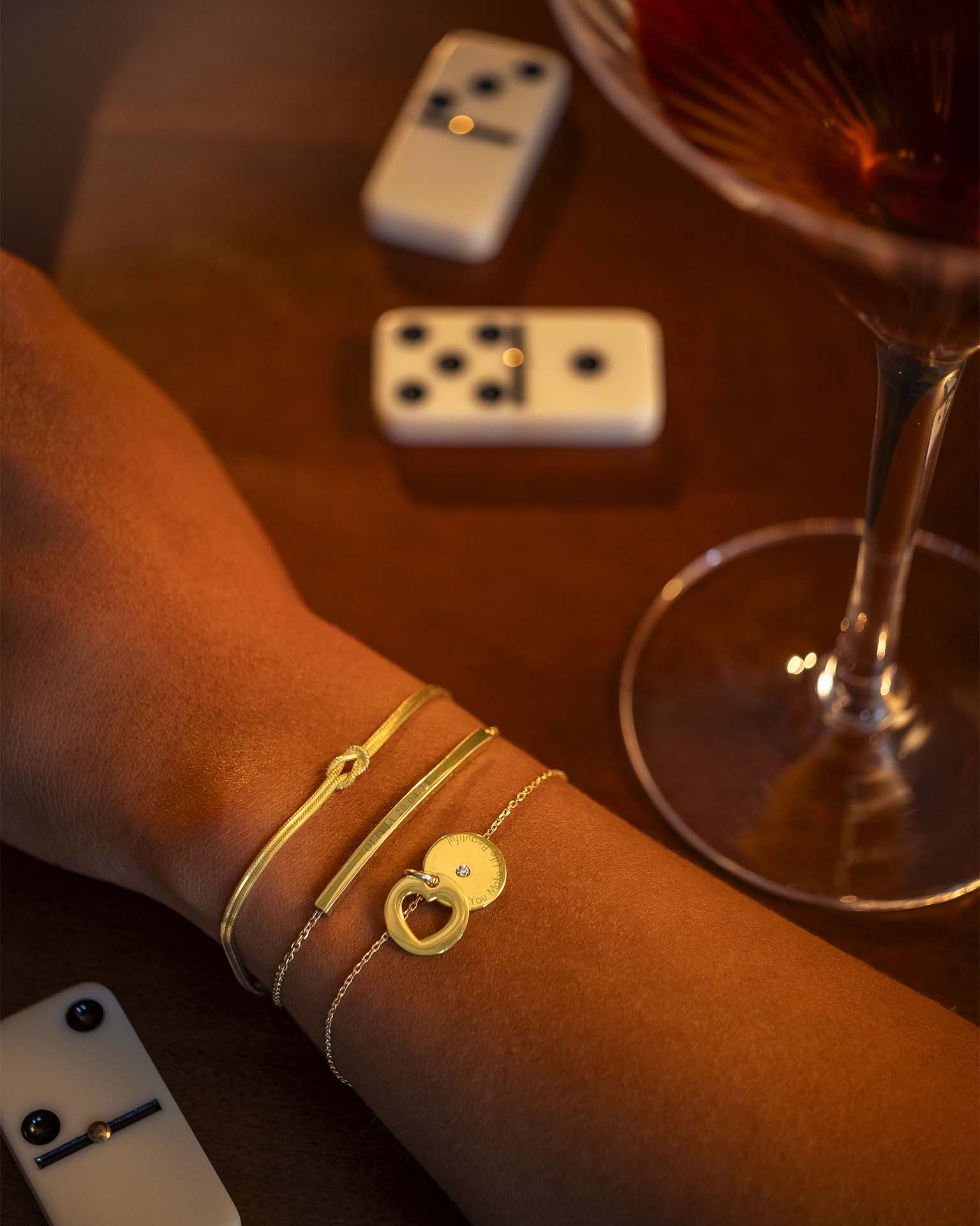 Gold bracelets on a wrist with a glass of red wine and dominoes in the background