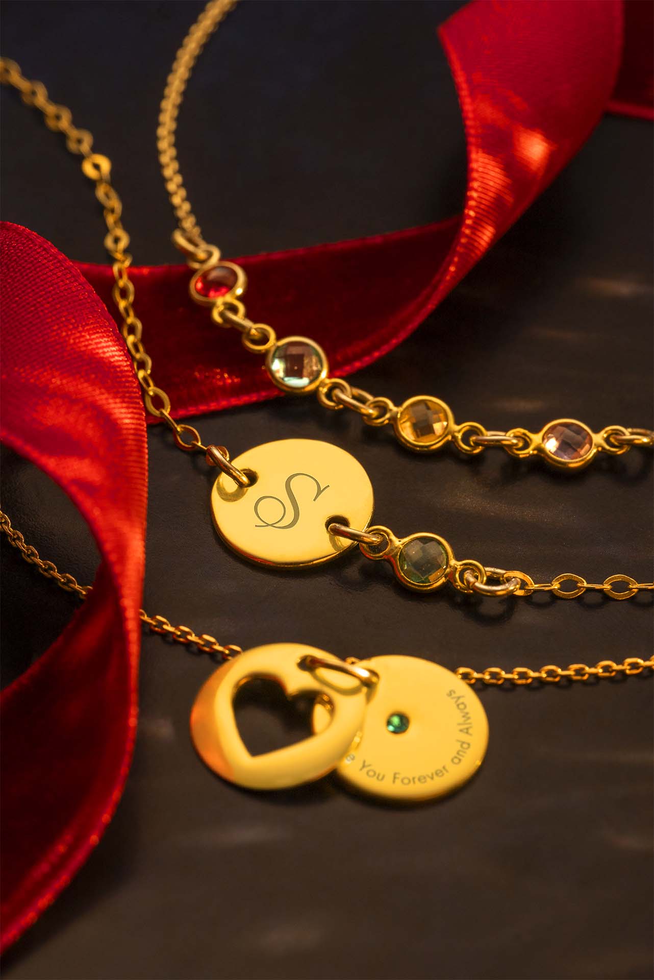 Gold necklaces with heart and round pendants on a dark surface with red ribbons.