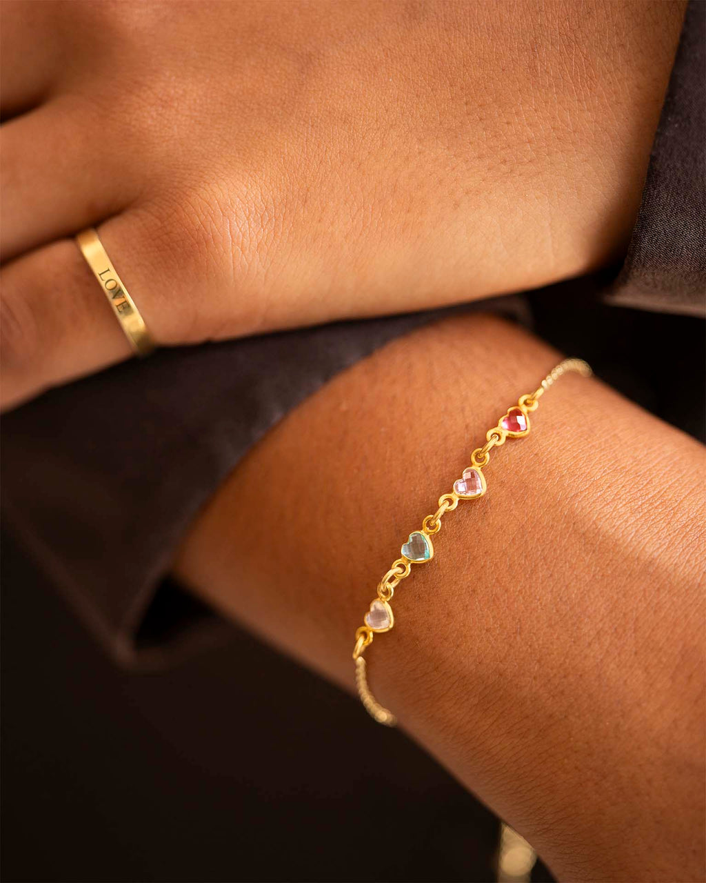 Gold bracelet with heart-shaped gemstones on a wrist, with a gold ring on a finger. | Christmas