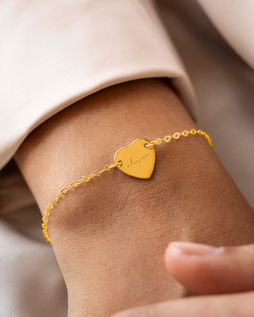 Gold bracelet with a heart-shaped tag on a wrist against a neutral background