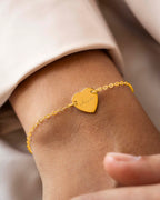 Gold bracelet with a heart-shaped tag on a wrist against a neutral background