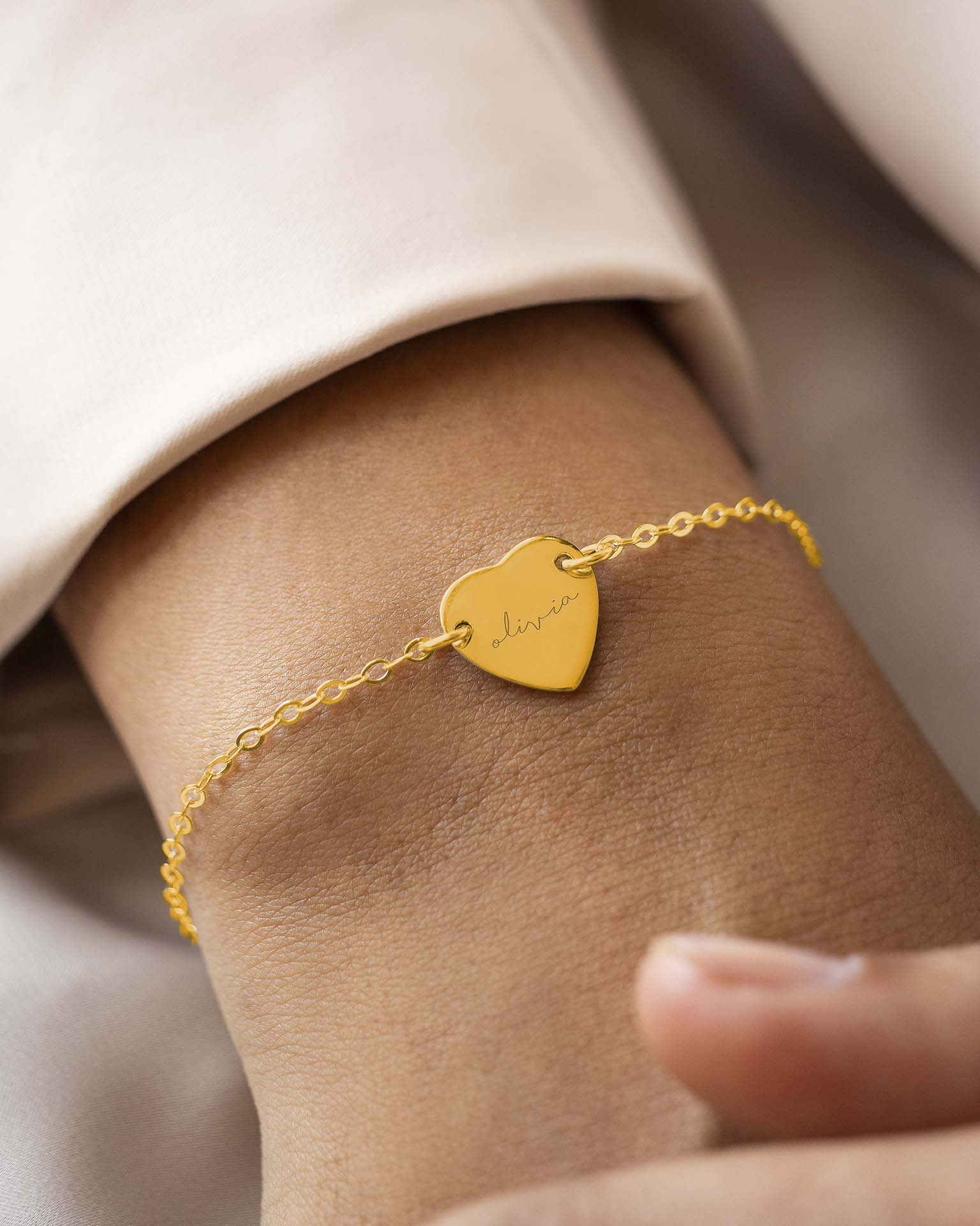 Gold bracelet with a heart-shaped tag on a wrist against a neutral background
