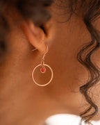 Close-up of a gold hoop earring with a red gemstone worn by a person.