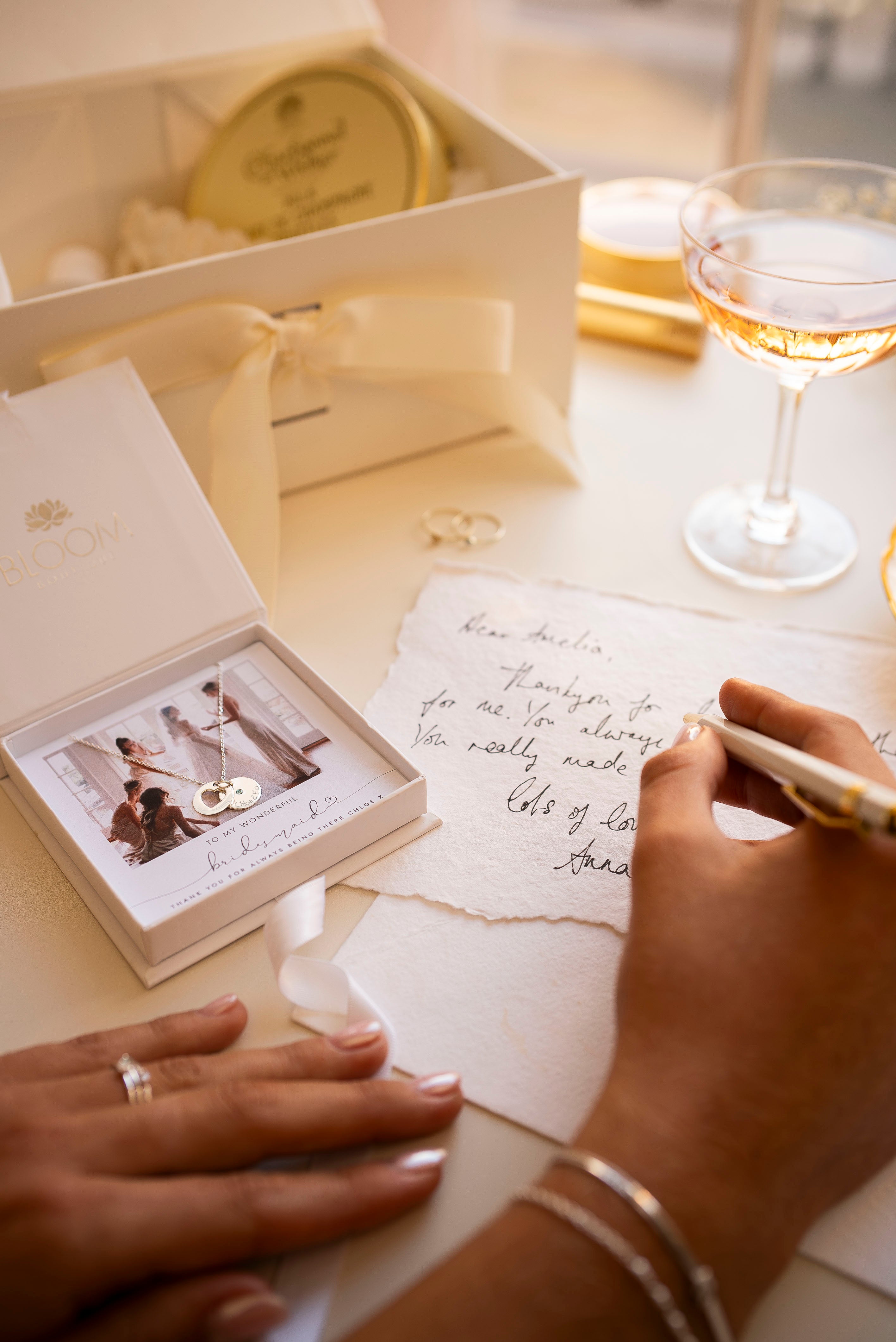 Person writing a letter with a box of chocolates and a glass of wine in the background