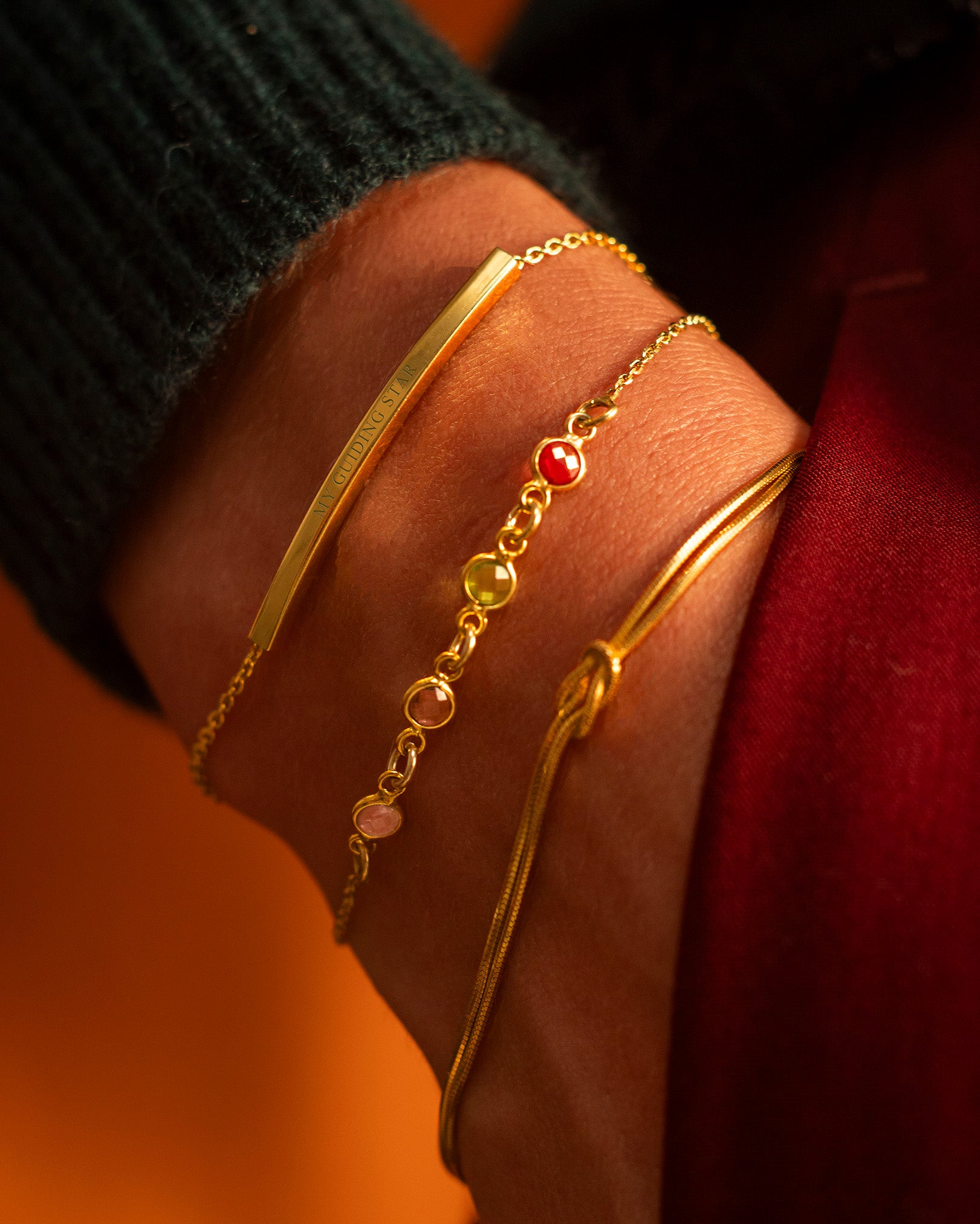 Close-up of a hand wearing gold bracelets with a red gemstone on an orange background